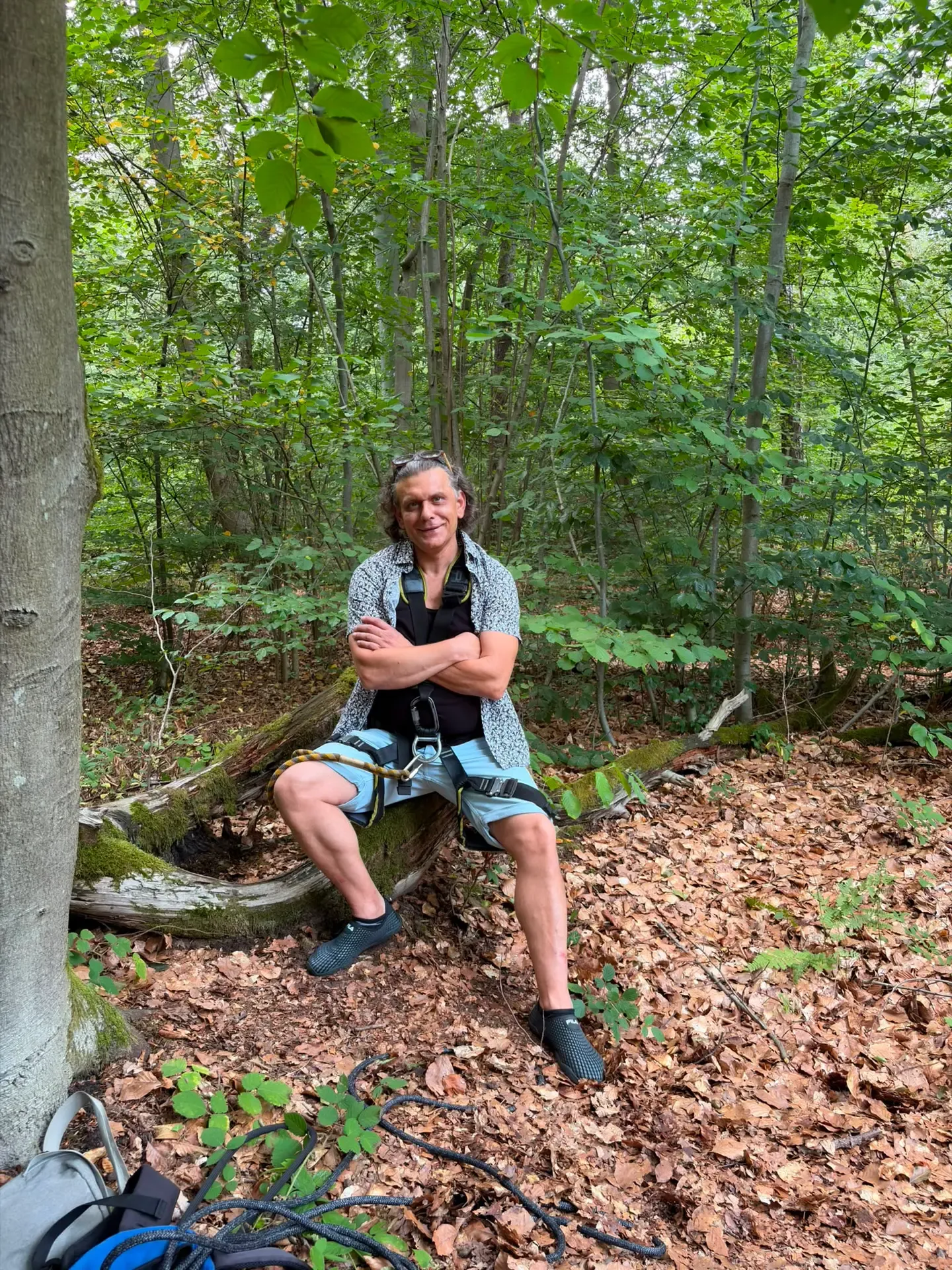 Sven Schwind sitzt lächelnd mit verschränkten Armen und Klettergurt auf einem Baumstamm im Wald